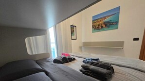Une chambre à coucher de taille modeste, avec deux lits, une fenêtre avec des rideaux, un tableau accroché au mur et une petite étagère.
