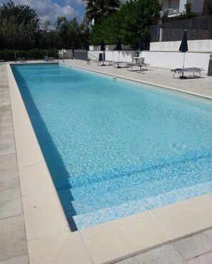 A rectangular swimming pool with clear blue water, surrounded by a tiled deck and shaded by umbrellas. - Apartment with swimming pool and large terrace a few steps from the sea, pets allowed (Vasto)