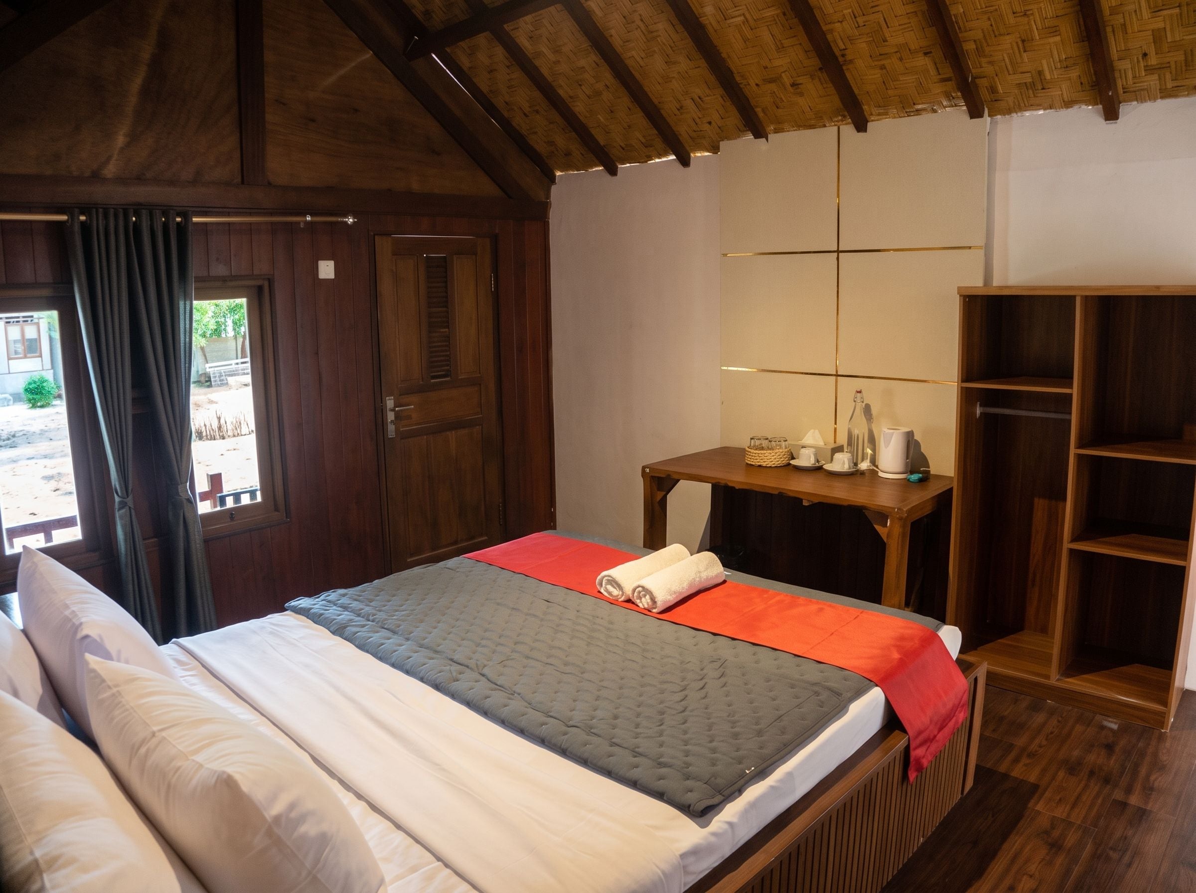 A room with a bed, a wooden wardrobe, a small table with a lamp, and a window with curtains.