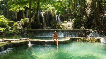 Eine Person sitzt in einem klaren Wasserbecken mit einem Wasserfall im Hintergrund.