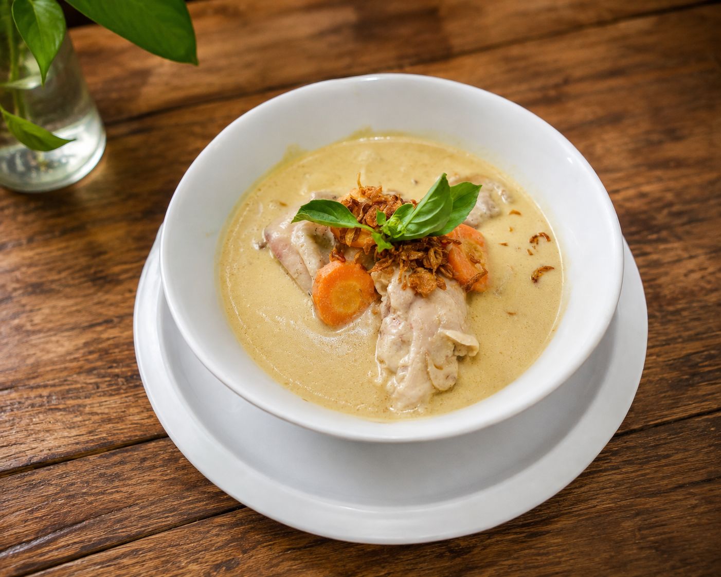 A bowl of soup with chicken, carrots, and a garnish on a wooden table.