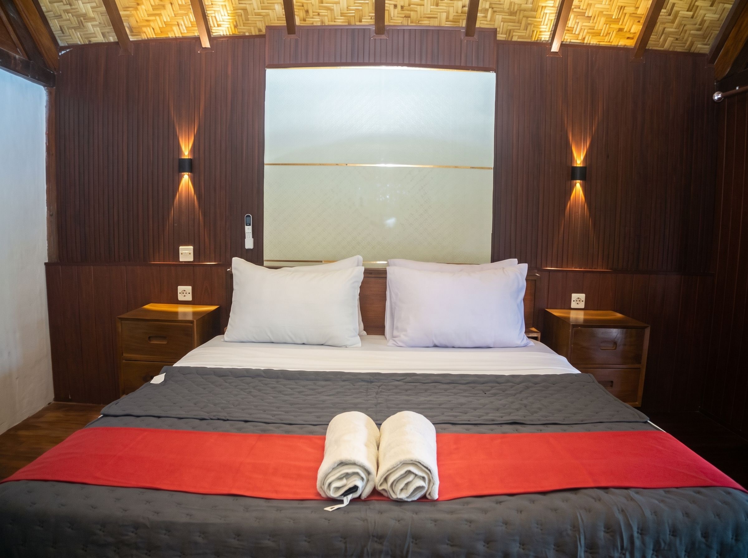 A hotel room with a large bed, two bedside tables, a nightstand, a wall-mounted light fixture, and a window.