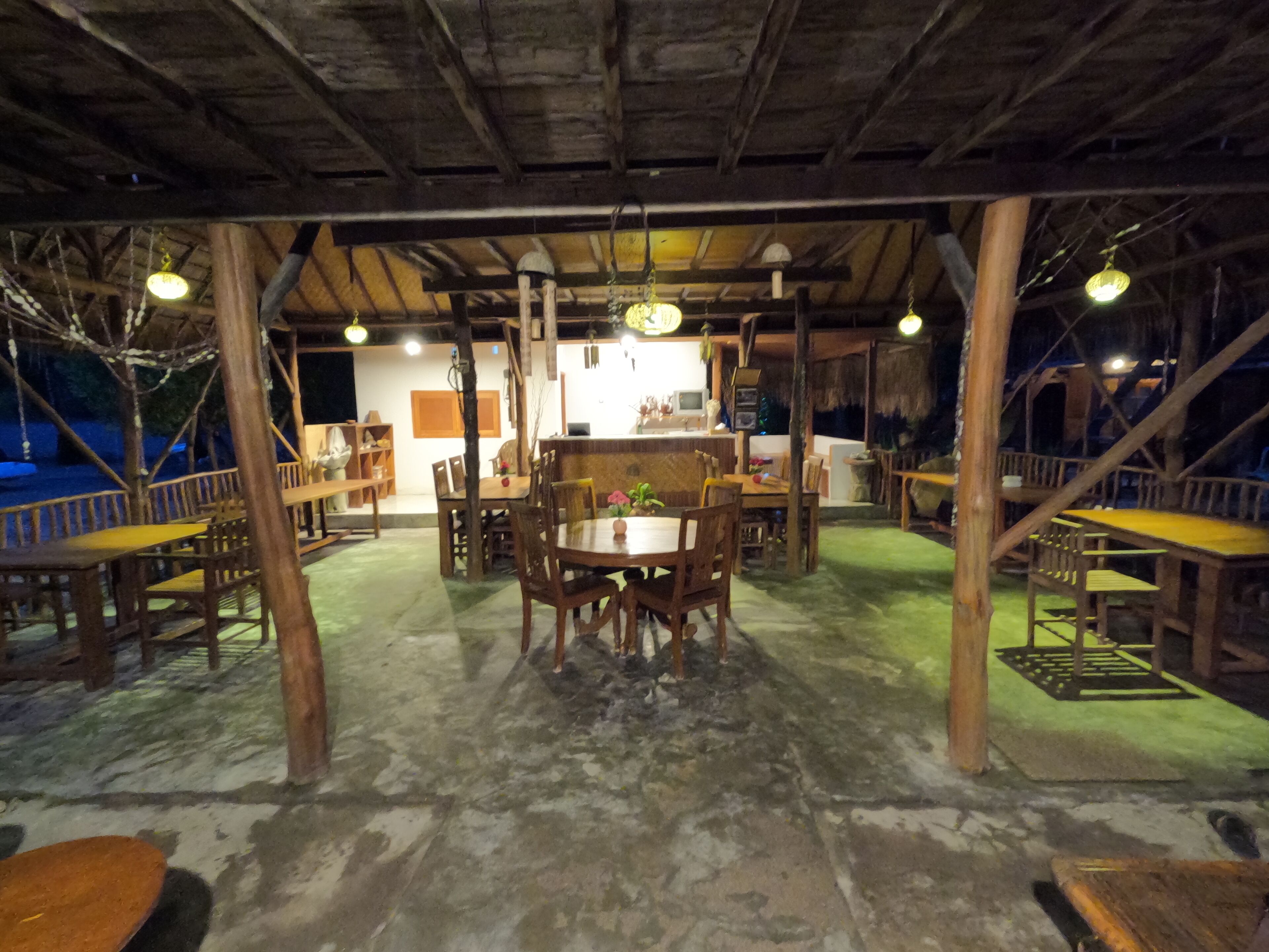 An outdoor wooden dining area with tables and chairs, a central table with a vase of flowers, and a thatched roof structure.