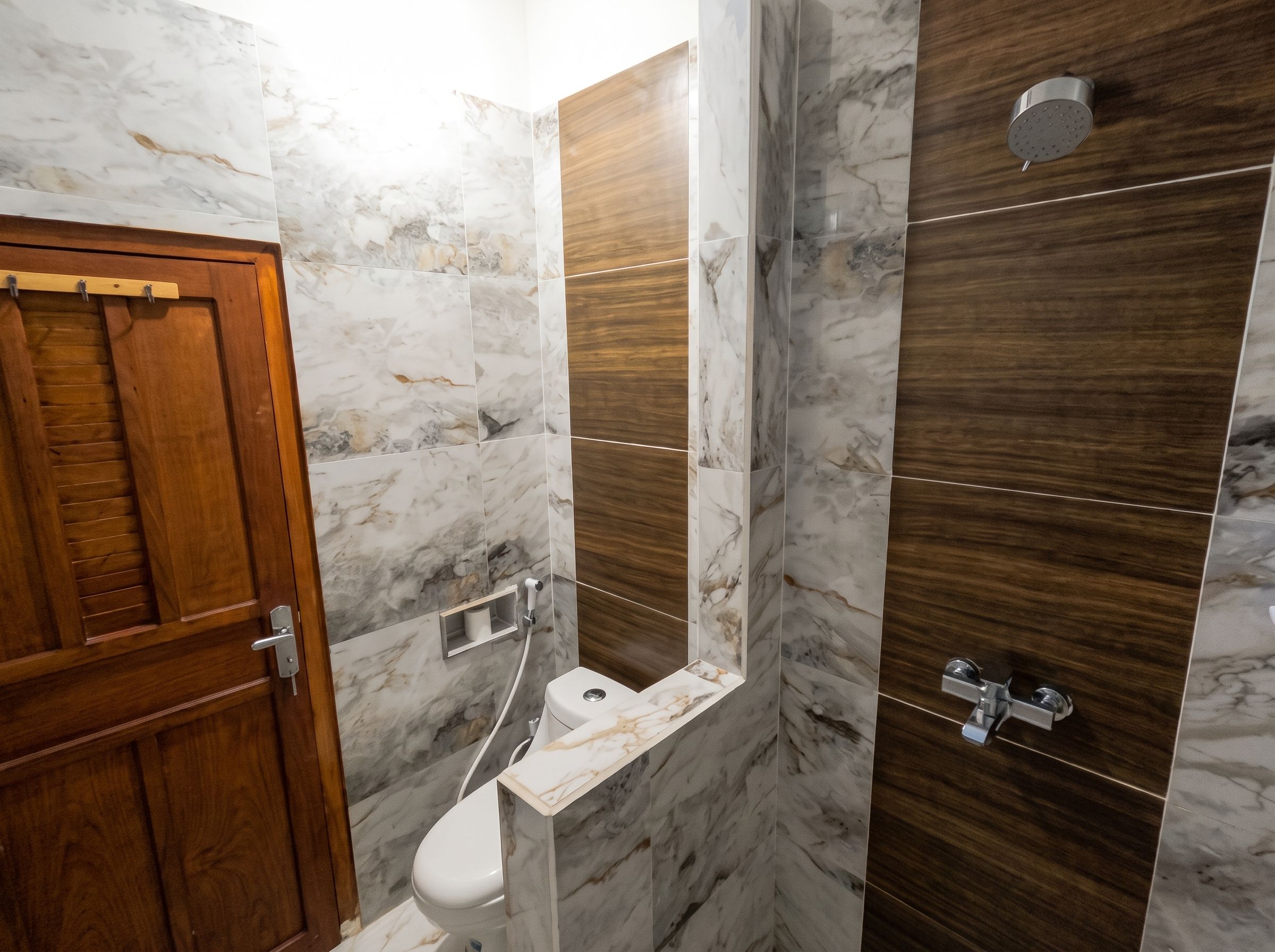A bathroom with a wooden door, a toilet, a shower with a wooden panel, and a marble wall.