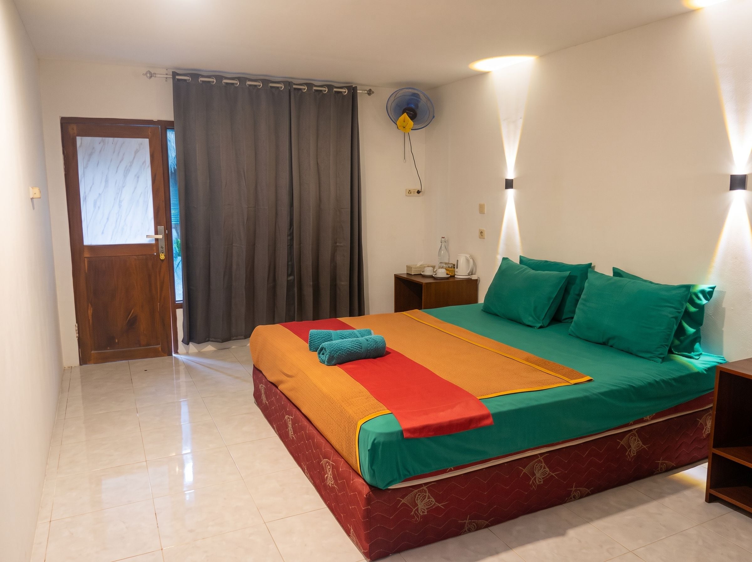 A hotel room with a bed, a nightstand, a fan, and a door.