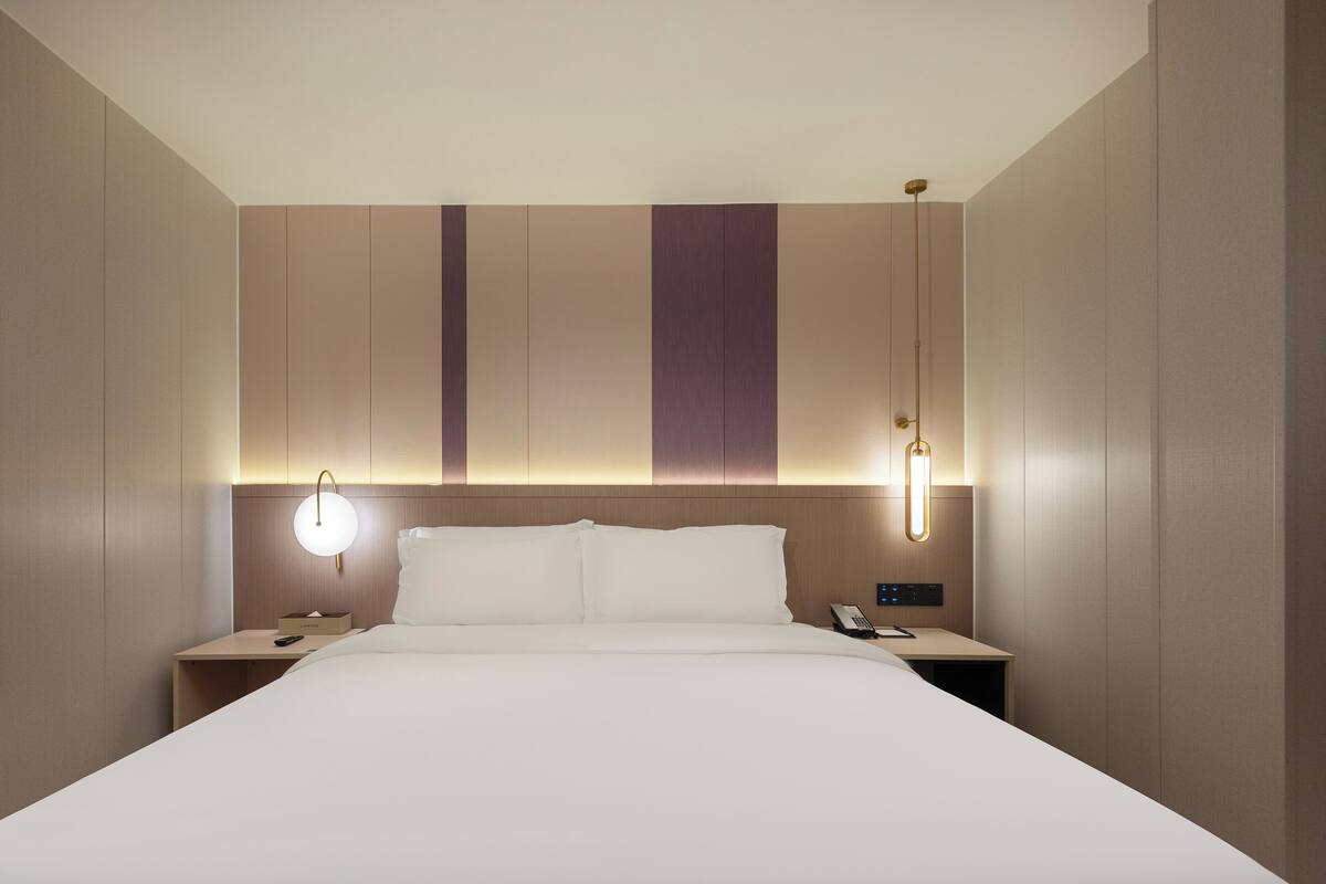 Una habitación de hotel con una cama grande, lámparas de noche, una mesita de noche y un panel de control en la pared.