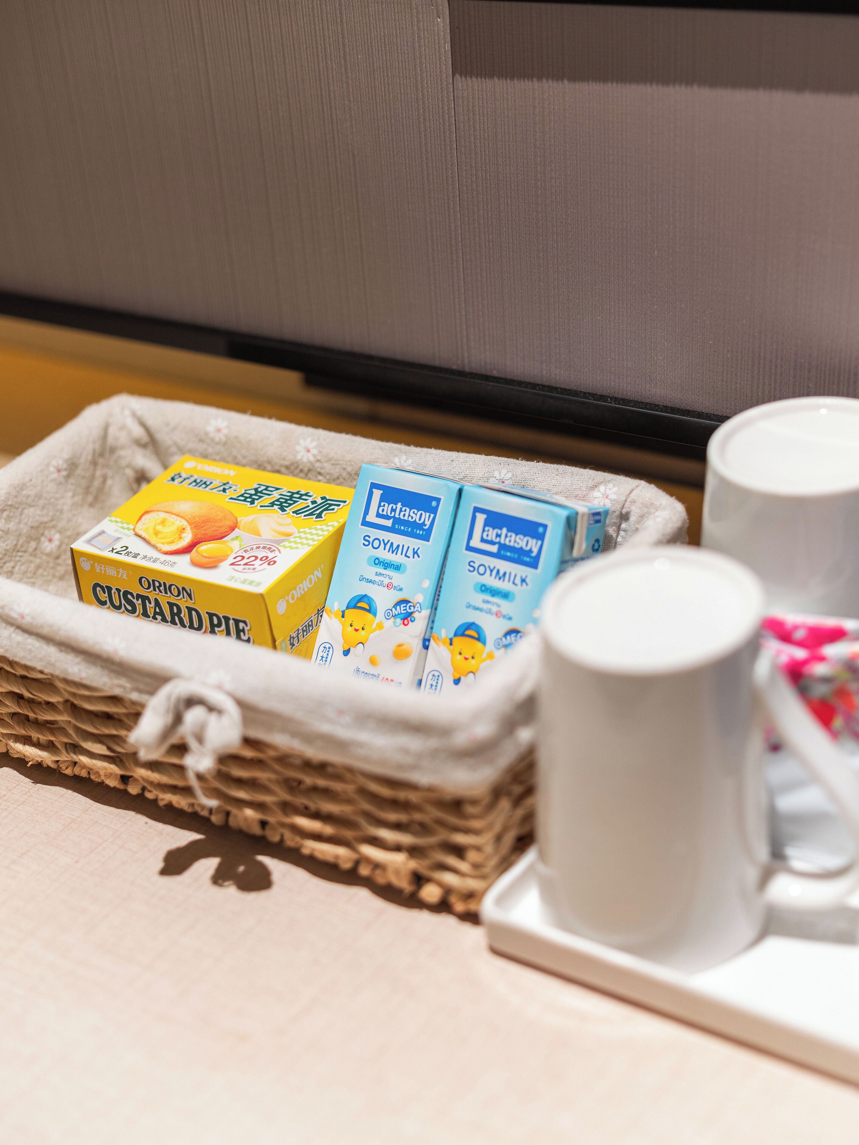 Un panier en osier contenant des boîtes de tarte à la crème Orion et de lait de soja Lactasoy, ainsi que deux tasses blanches posées sur un plateau.
