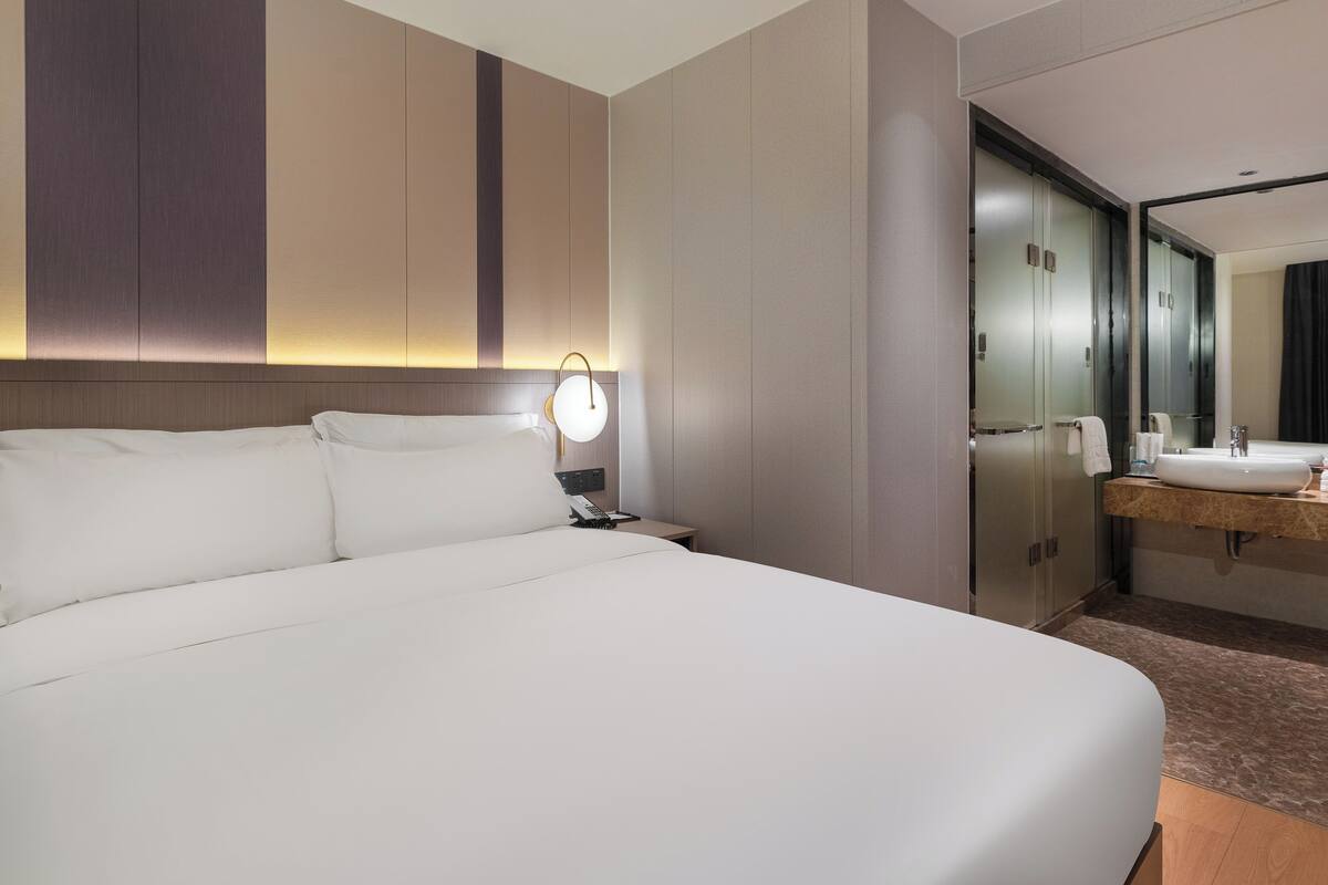 Habitación de hotel con una cama grande, una mesita de noche con lámpara, un espejo y un baño con lavamanos y espejo.