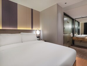 Habitación de hotel con una cama grande, una mesita de noche con lámpara, un espejo y un baño con lavamanos y espejo.