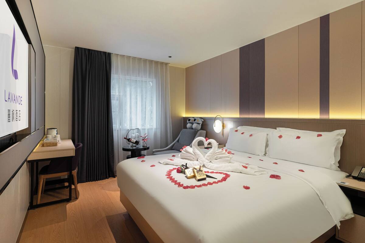 Habitación de hotel con una cama bien hecha, un escritorio con silla, un ventanal con cortinas y un televisor montado en la pared.