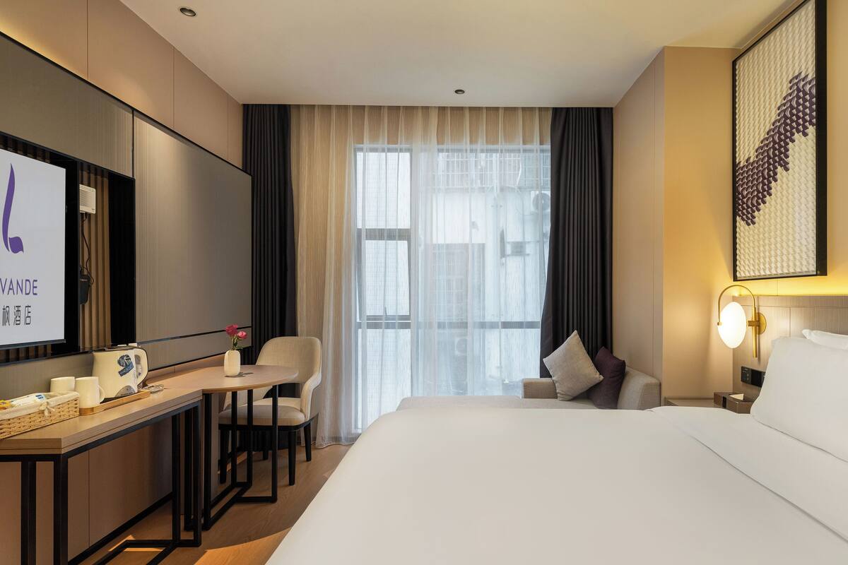 Une chambre d’hôtel comprenant un lit, un bureau avec une chaise, une télévision et une grande fenêtre avec des rideaux.