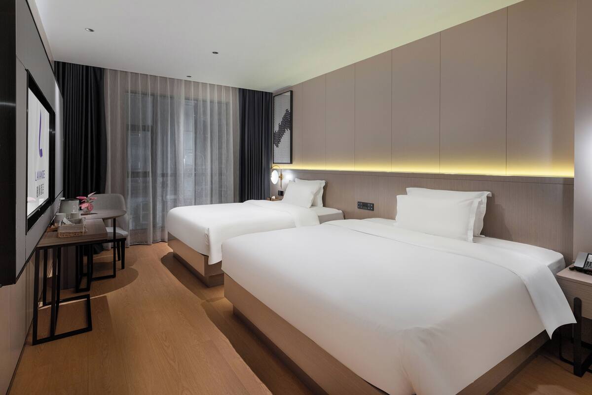 Une chambre d’hôtel avec deux lits, un bureau, une chaise et une grande fenêtre avec des rideaux.