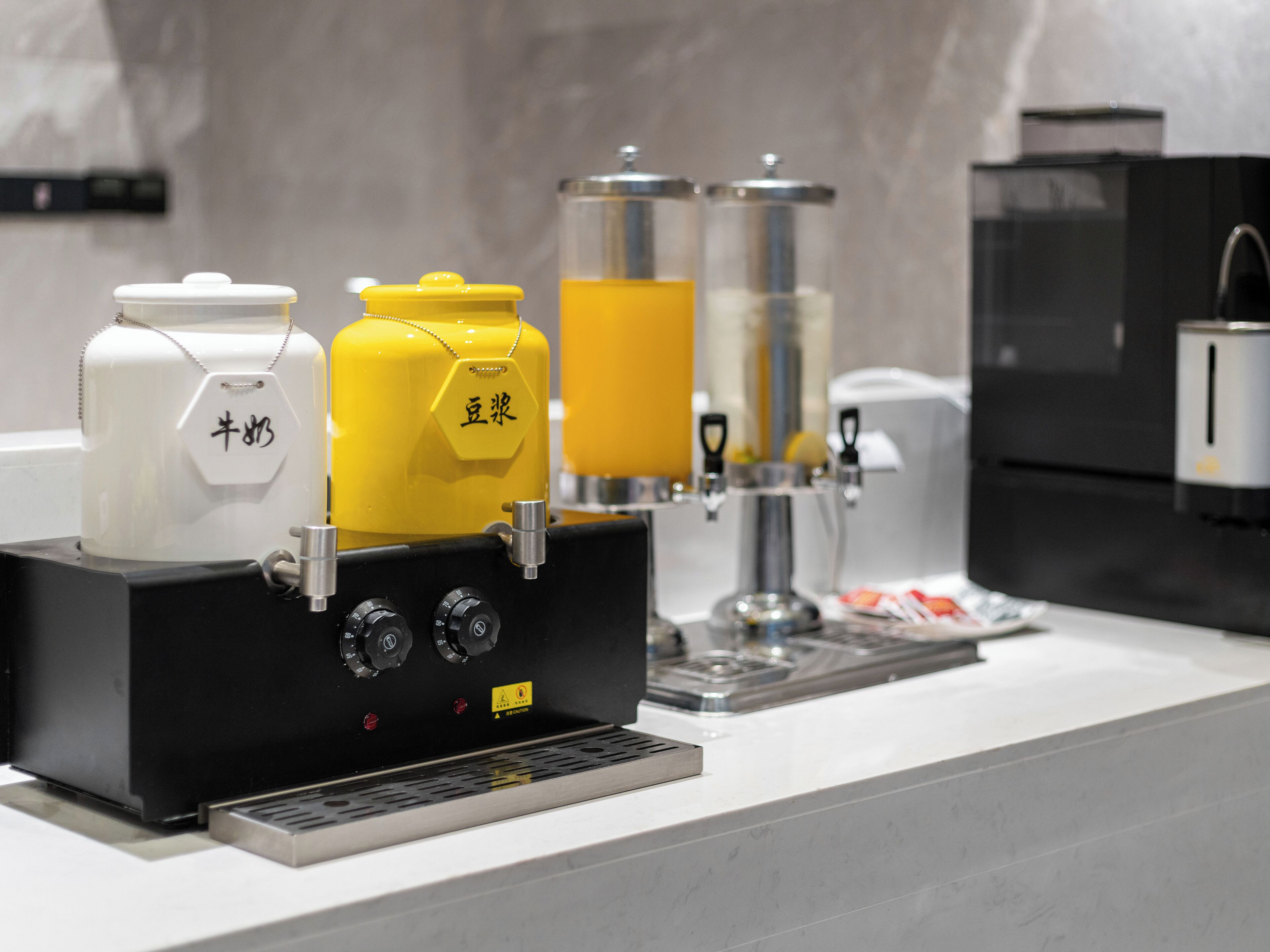 Una máquina de café con base negra y recipientes amarillos y blancos, un dispensador de café y un refrigerador al fondo.