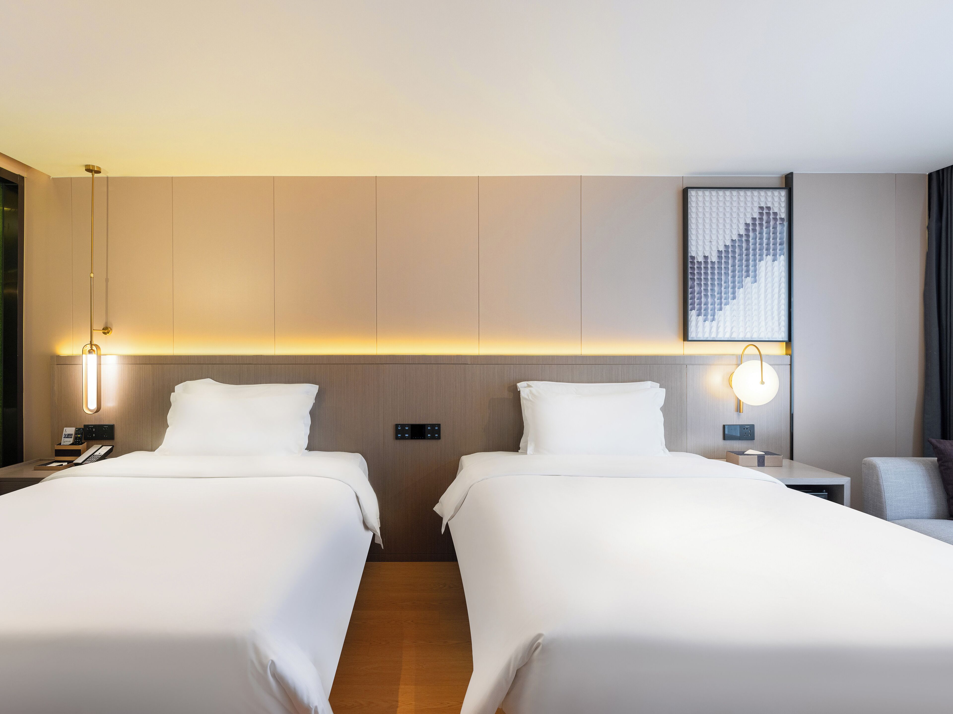 Une chambre d’hôtel avec deux lits, une table de chevet avec une lampe, un panneau de commande fixé au mur et une œuvre d’art encadrée au-dessus des lits.