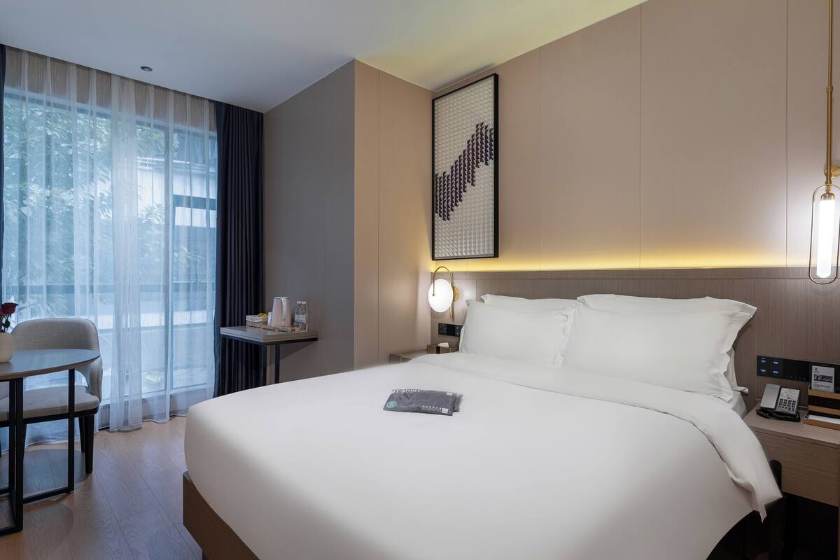 Une chambre d’hôtel avec un grand lit, une table de chevet avec une lampe, une chaise et une vue sur les arbres à travers de fines rideaux.