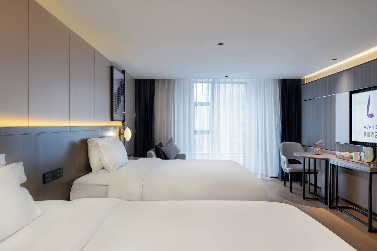 Une chambre d’hôtel avec deux lits, un bureau avec une chaise, une télévision et une grande fenêtre avec des rideaux.