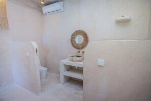 A bathroom with a toilet, a sink with a decorative bowl, a mirror, and an air conditioning unit on the wall.