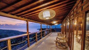 Un balcon en bois avec des fauteuils en osier, une vue sur la mer et un coucher de soleil.