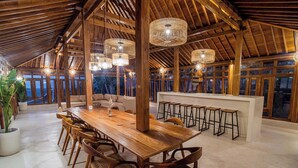 Un vaste espace dédié à la salle à manger, avec une longue table en bois et des chaises, un bar avec des tabourets, et de grandes fenêtres offrant une vue sur l’extérieur.