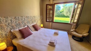 Una cama bien hecha con almohadas, una lámpara de noche y una silla en una habitación con ventana que ofrece vistas al verdor.
