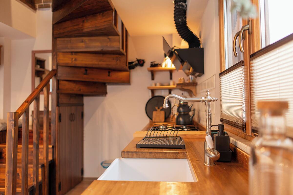 Una cocina con armarios de madera, un lavabo blanco, una tetera y una ventana con persianas.
