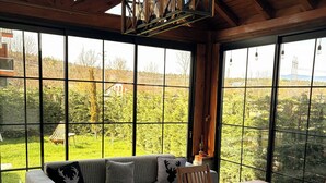A spacious room with a wooden ceiling, large windows, a grey sofa, a wooden dining table, and a hanging light fixture. - Abant Cozy Homes (Bolu)