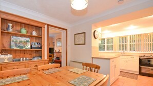 Un coin repas avec une table et des chaises en bois, une cuisine avec des meubles blancs et une fenêtre équipée de stores.