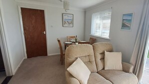 A cozy living room with a beige sofa, a dining table with chairs, a wooden door, and a window with blinds. - Brambles Farm Bungalow (Freshwater)