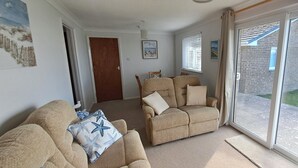 A living room with beige sofas, a wooden door, and a view of a brick building through a glass door. - Brambles Farm Bungalow (Freshwater)