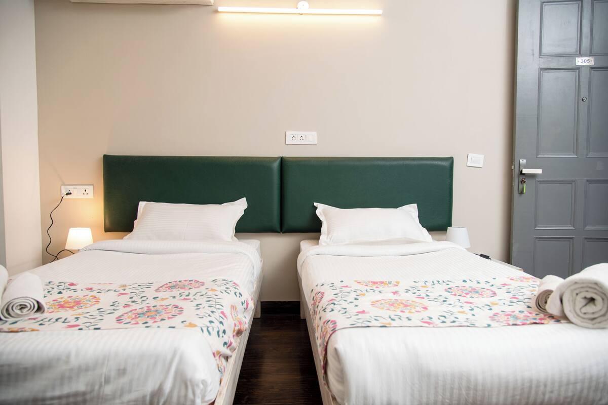 Habitación de hotel con dos camas individuales, cabecera verde, ropa de cama blanca y una puerta con número.