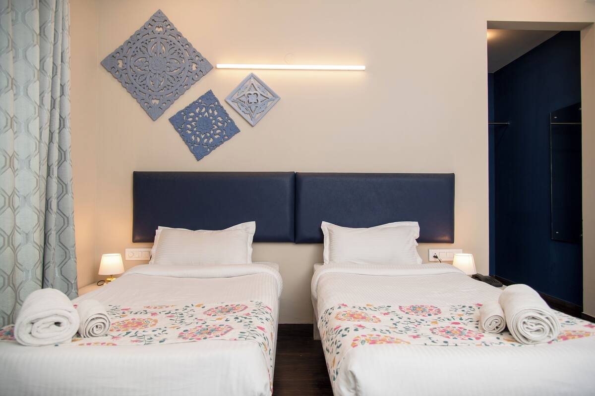 Habitación de hotel con dos camas, cabecero oscuro, decoración mural y mesita de noche con lámpara.