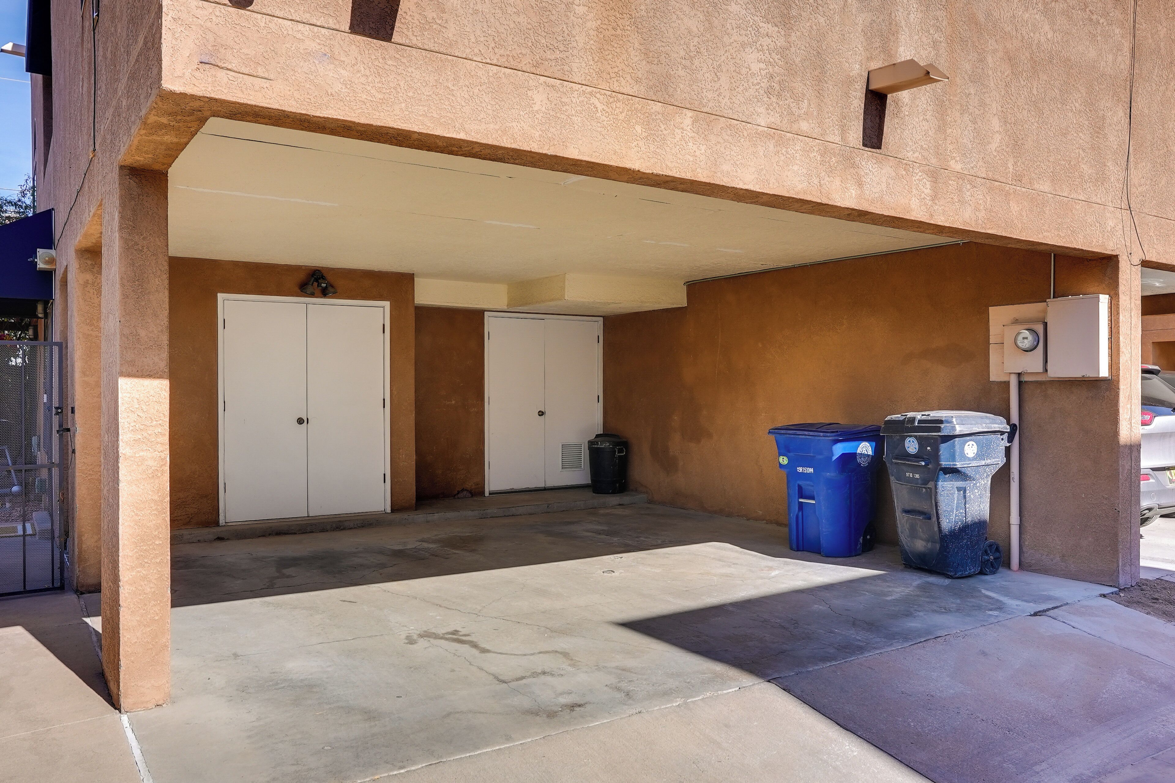 A covered parking area with two white doors, a trash bin, and two blue trash bins.