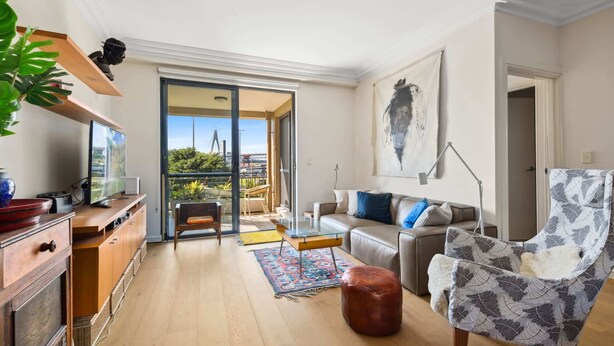 Un salon moderne avec un canapé, un pouf et un fauteuil orné d’un motif de feuilles. On y trouve un téléviseur posé sur un meuble en bois, une plante et une vue sur l’extérieur grâce à une porte vitrée.