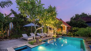 A swimming pool with lounge chairs and umbrellas, surrounded by lush greenery and a building with a red roof. - Le Biu Garden View (Lembongan Island)