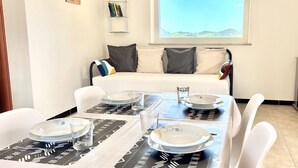 A dining area with a table set for four, a sofa with cushions, a bookshelf, and a window with a view. - Residence Sole - Apartment 2 (Tertenia)