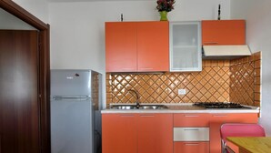 Keittiössä on oranssit kaapit, ruostumattomasta teräksestä valmistettu jääkaappi ja laatoitettu välitila.