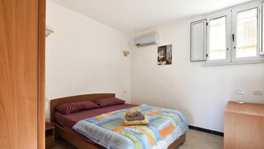Une chambre à coucher comprenant un lit, une table de chevet en bois, un appareil de climatisation, une fenêtre et une armoire en bois.