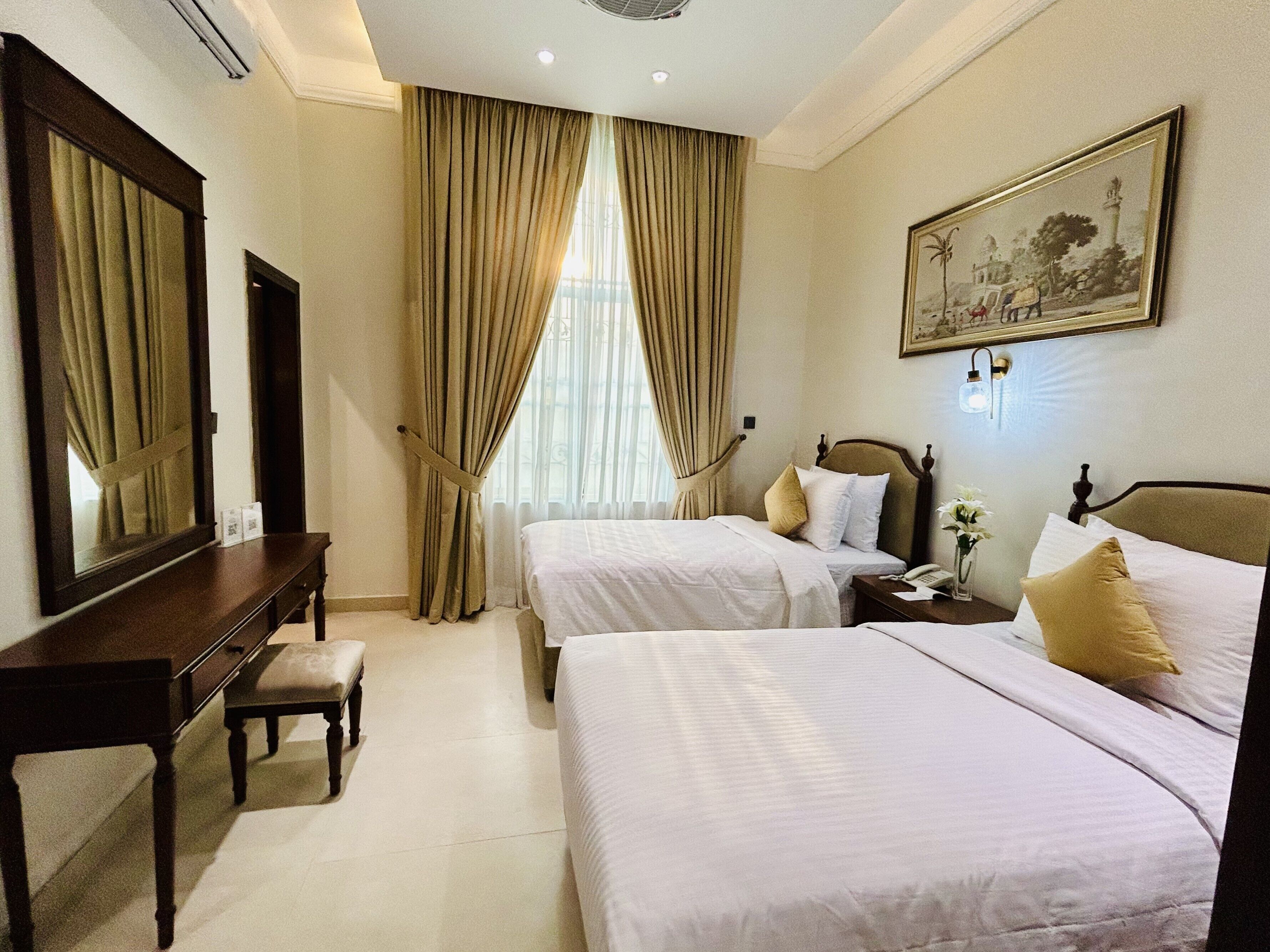 A hotel room with two beds, a desk, a chair, a nightstand, and a painting on the wall.