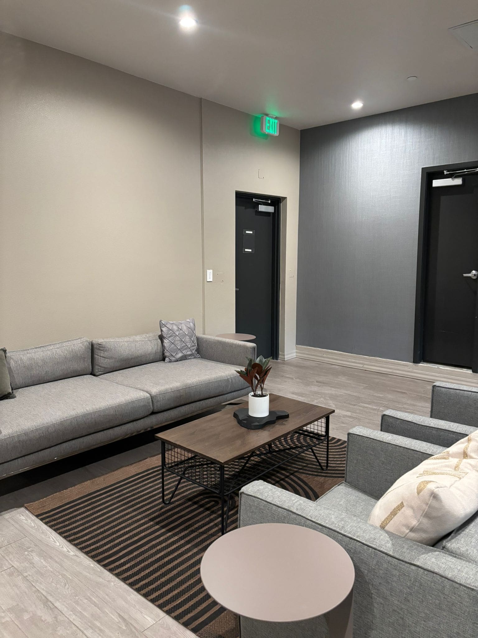 A modern lobby with a grey sofa, a coffee table, and a small round table. There are two doors and an exit sign on the wall.