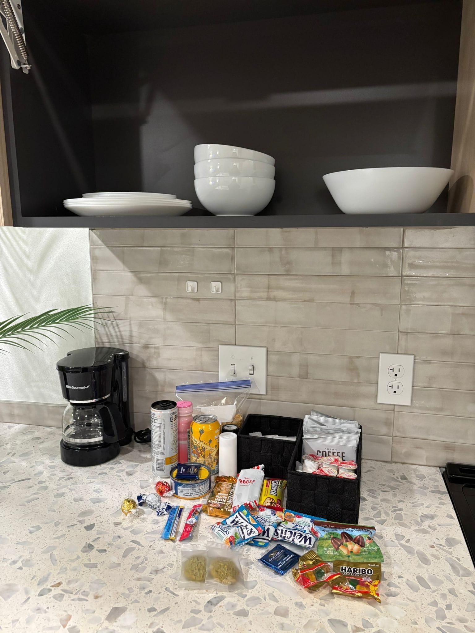 A kitchen counter with a coffee maker, various snacks, and a basket of newspapers.