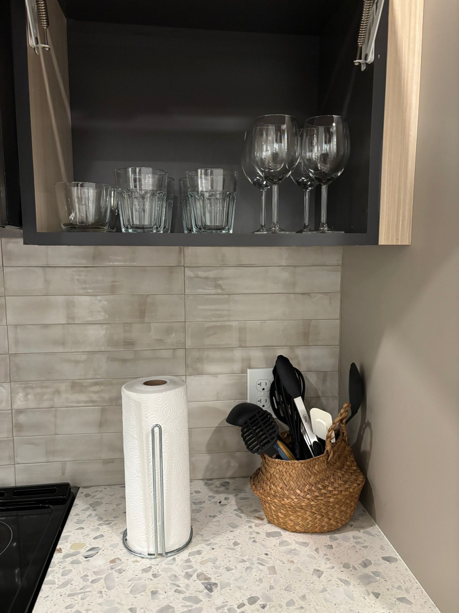 A kitchen with a countertop, a roll of paper towels, a woven basket with utensils, and a shelf with various glassware.