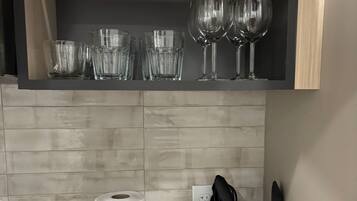 A kitchen with a countertop, a roll of paper towels, a woven basket with utensils, and a shelf with various glassware.