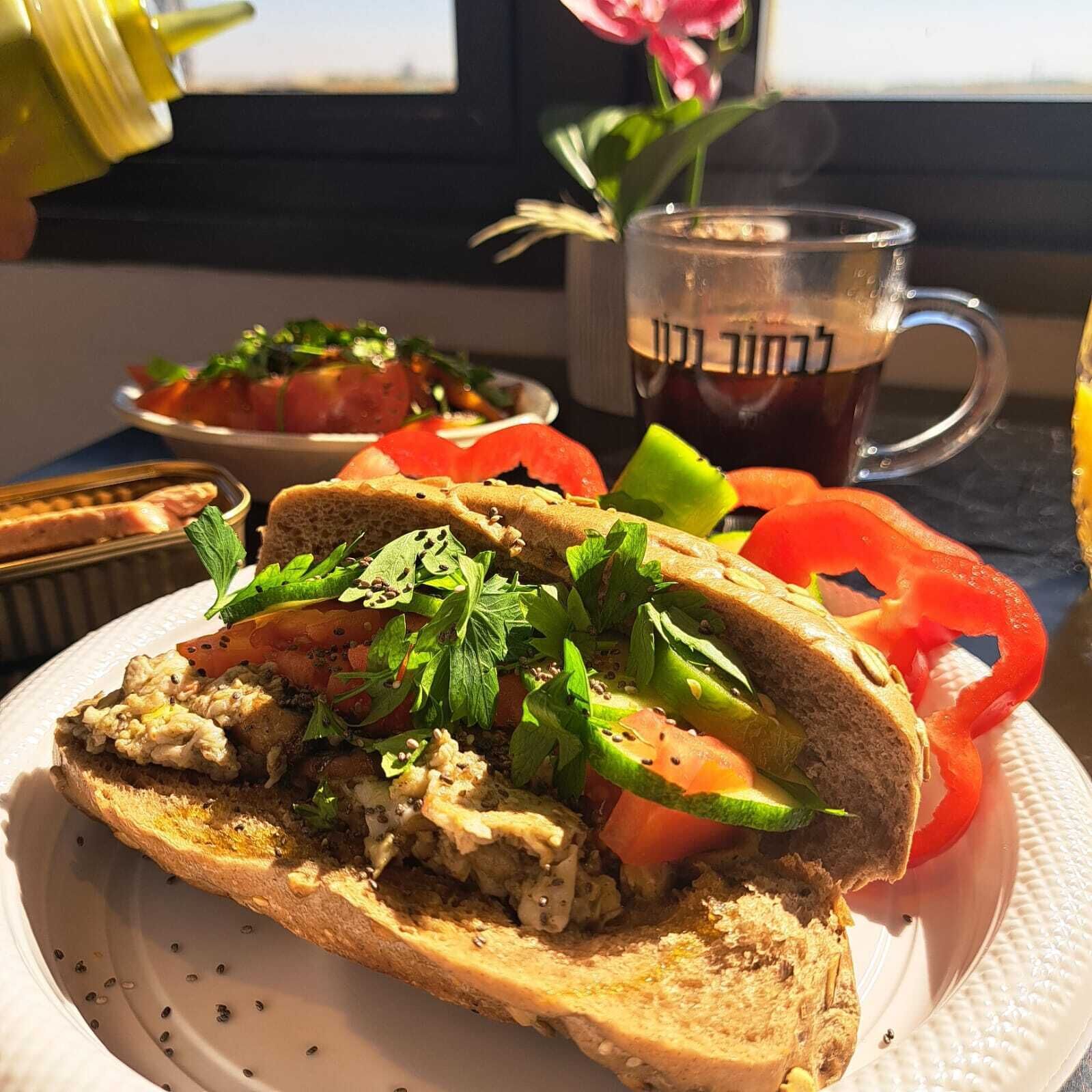 A sandwich with various toppings, a glass of tea, and a flower on a table.