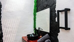 A bathroom with a stone wall, a mirror, a sink with a black basin, and a wooden shelf with a towel.