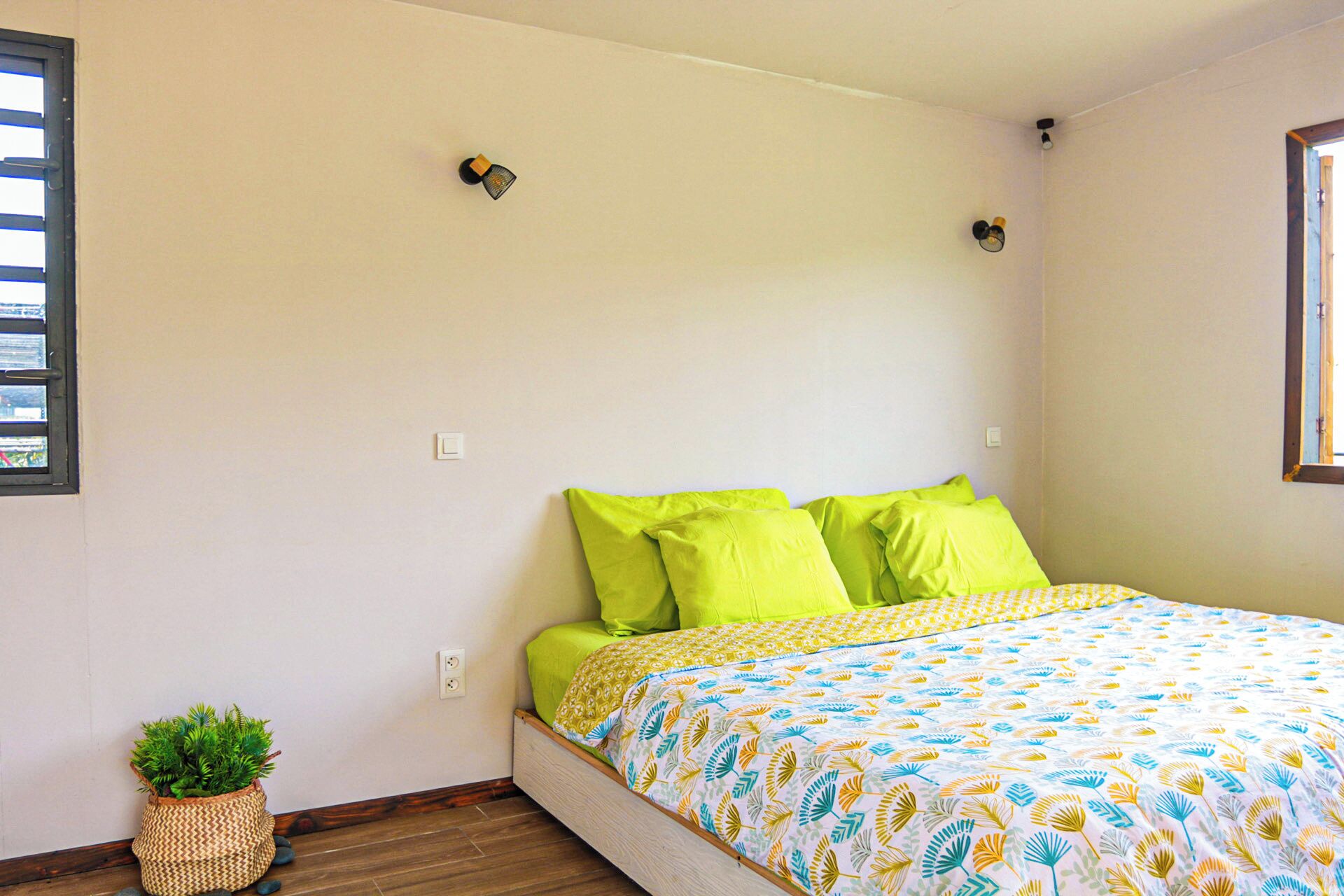 Une chambre à coucher avec un lit, des oreillers verts, une couvre-lit à motifs, une petite plante en pot et une fenêtre avec des stores.