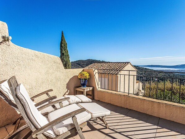 A balcony with two lounge chairs, a small table with a vase of flowers, and a view of the sea and hills.