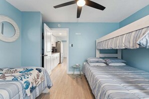 Une chambre avec deux lits, un ventilateur de plafond, une petite table et une vue sur la cuisine.
