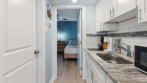 A narrow hallway leading to a bedroom with a bed, a kitchen with white cabinets and a sink, and a blue accent wall.