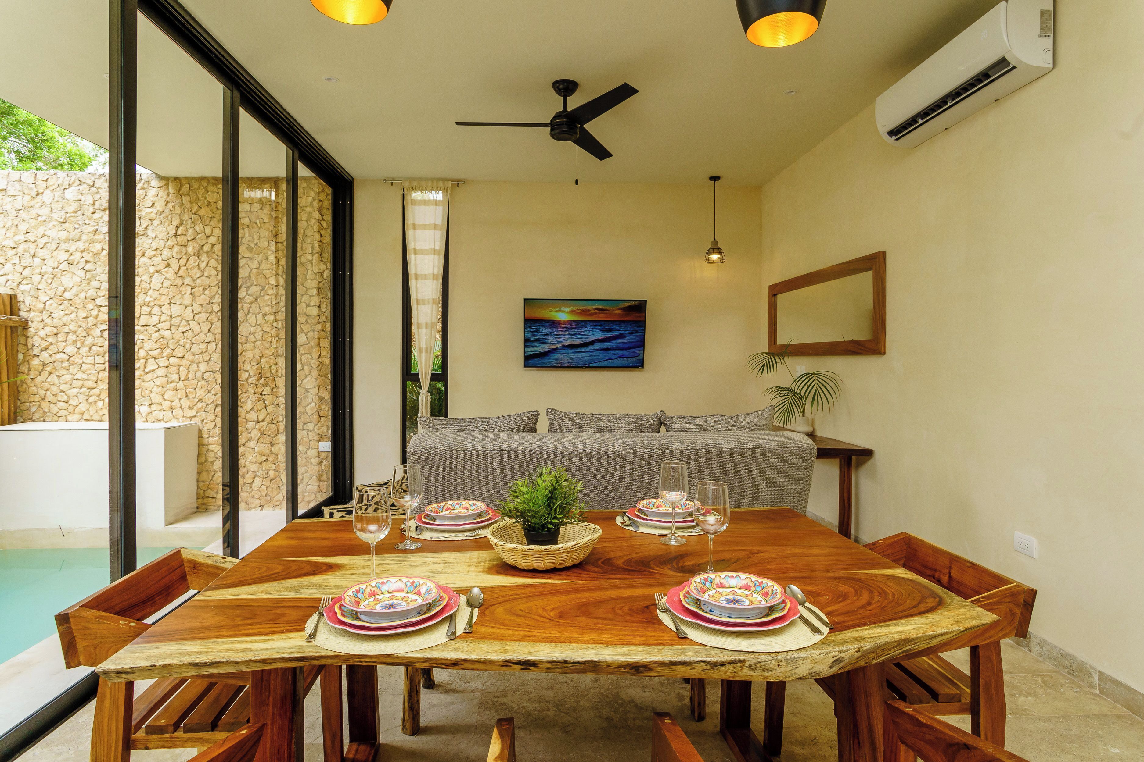 Une table à manger dressée avec des assiettes, des verres et des couverts, entourée de chaises en bois, et une piscine visible à travers des portes vitrées.
