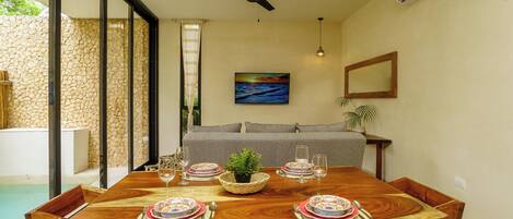 Une table à manger dressée avec des assiettes, des verres et des couverts, entourée de chaises en bois, et une piscine visible à travers des portes vitrées.